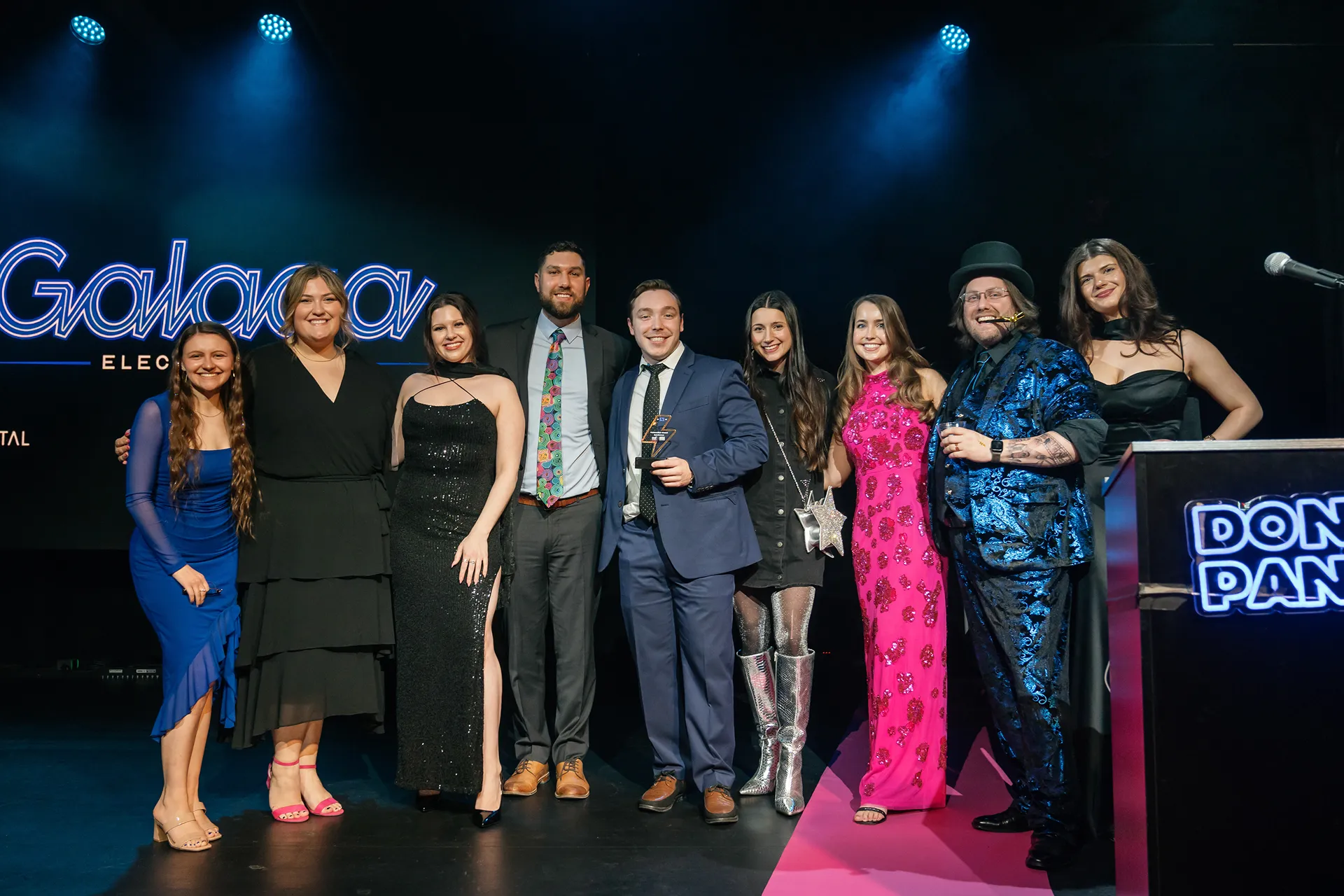 Group of eight well-dressed people posing on stage with a trophy under blue stage lights.