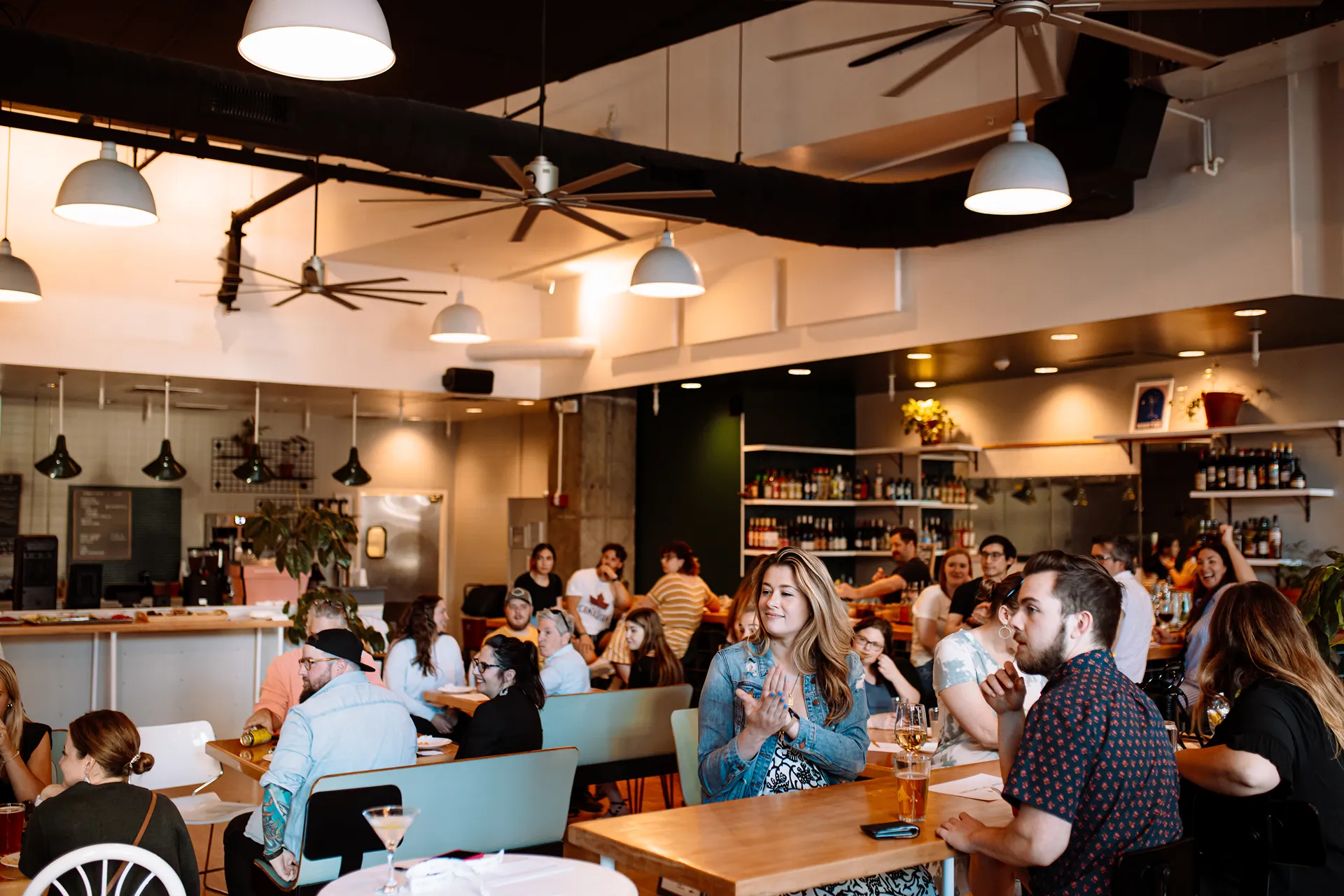 People socializing and seated at tables in a lively, modern bar or restaurant with pendant lights and ceiling fans.