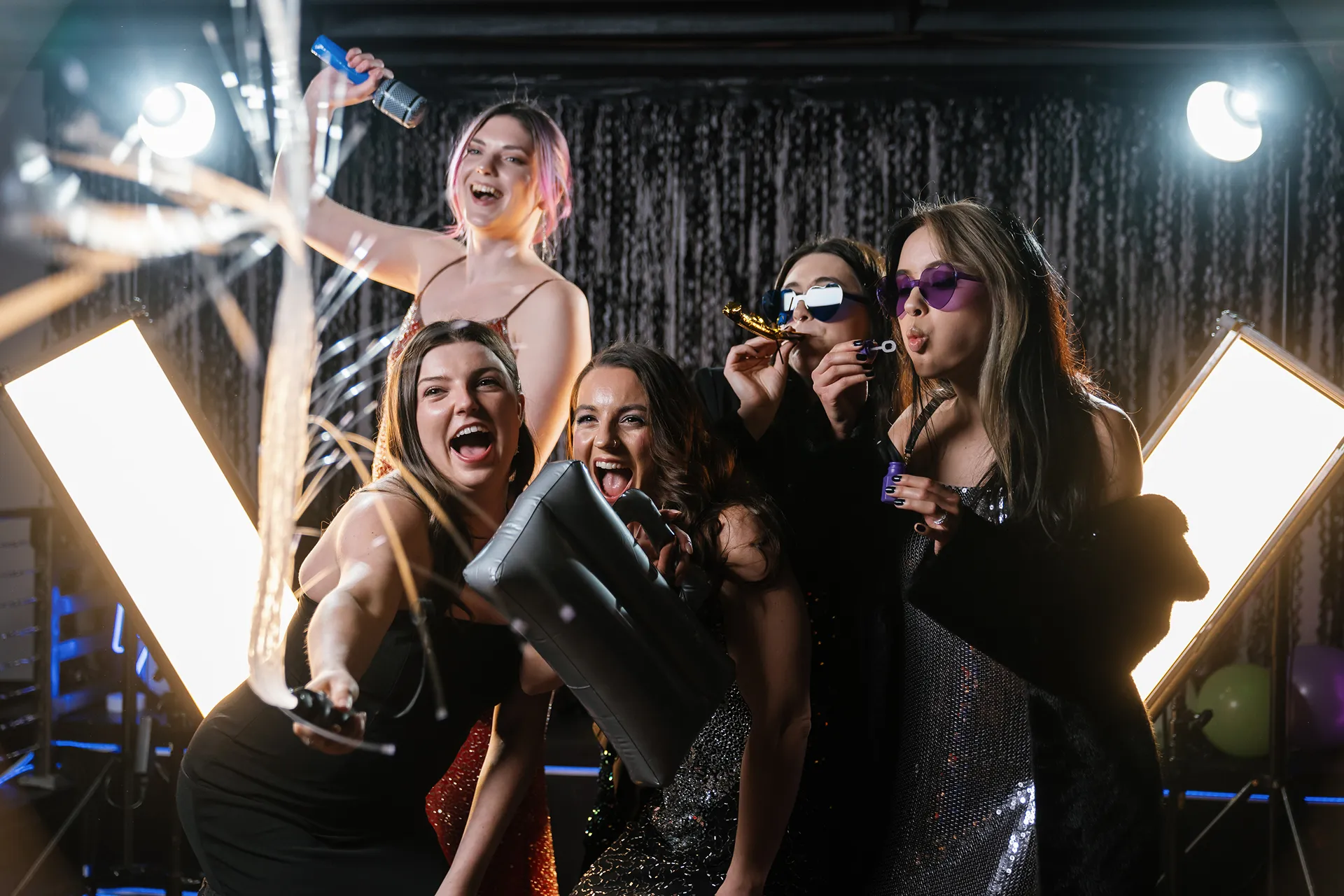 Group of five women dressed in party attire enjoying a celebration, with sparklers, inflatable props, and blowing bubbles.