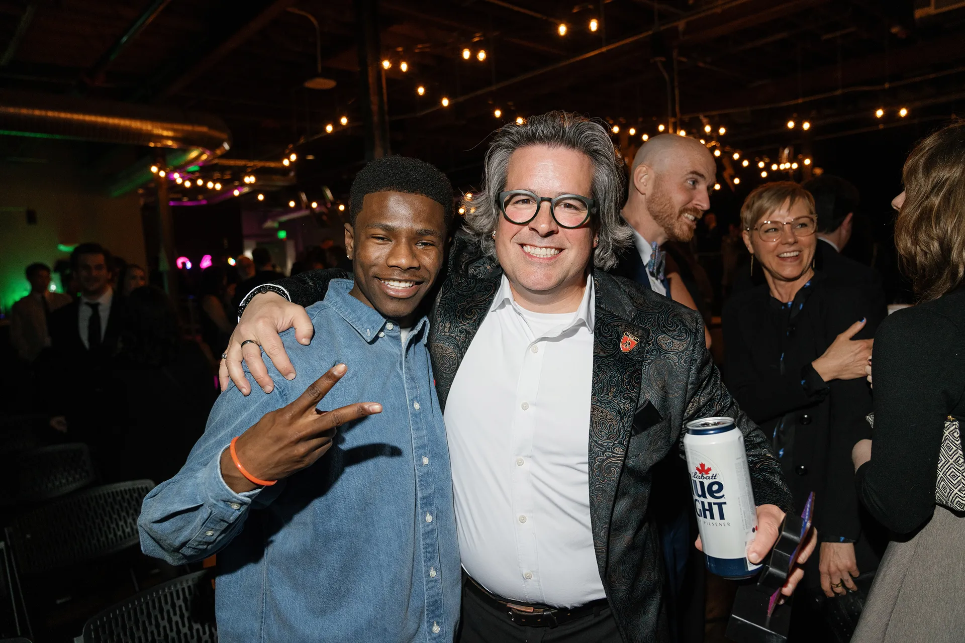 Two men smiling at a social event, one making a peace sign and the other holding a can of Labatt Blue Light beer.