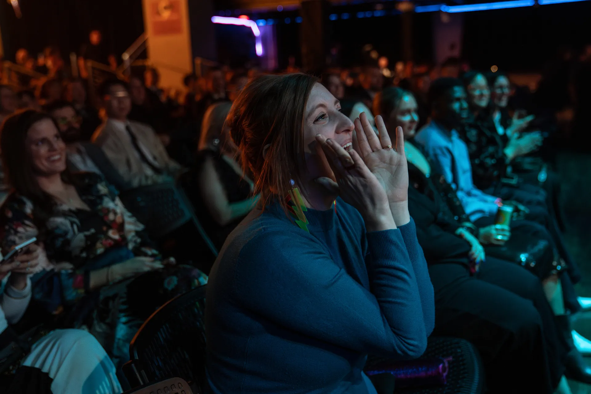 Woman in a blue sweater cheering and cupping hands around her mouth at an indoor event with seated audience.