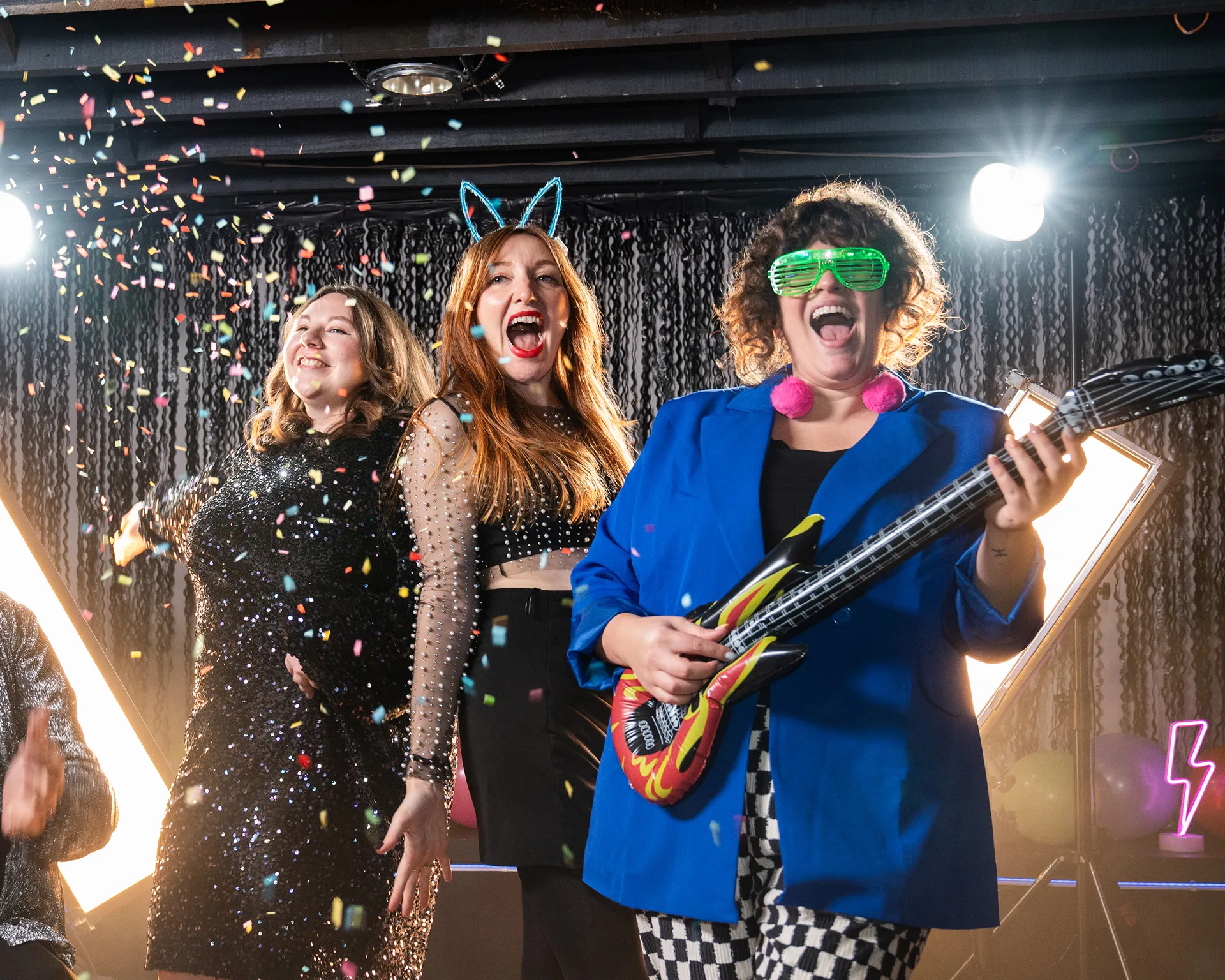 Three women celebrating with confetti; one plays a toy guitar wearing neon green shutter shades, one wears blue light-up bunny ears and another in a sparkly black dress.