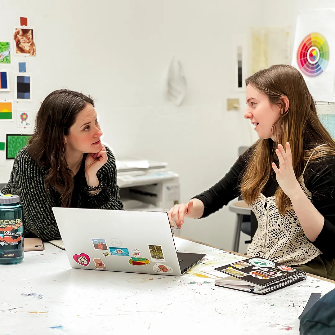 Two women engaged in a discussion across a white table with a laptop, notebook, and water bottle.