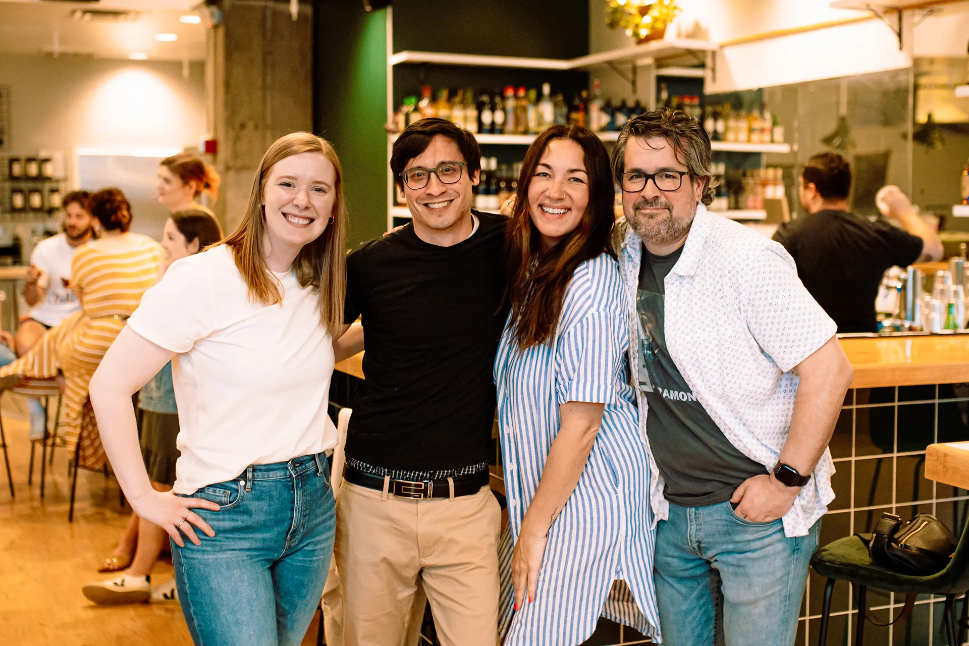 Four people smiling and posing together inside a bar or cafe with bottles on shelves in the background.
