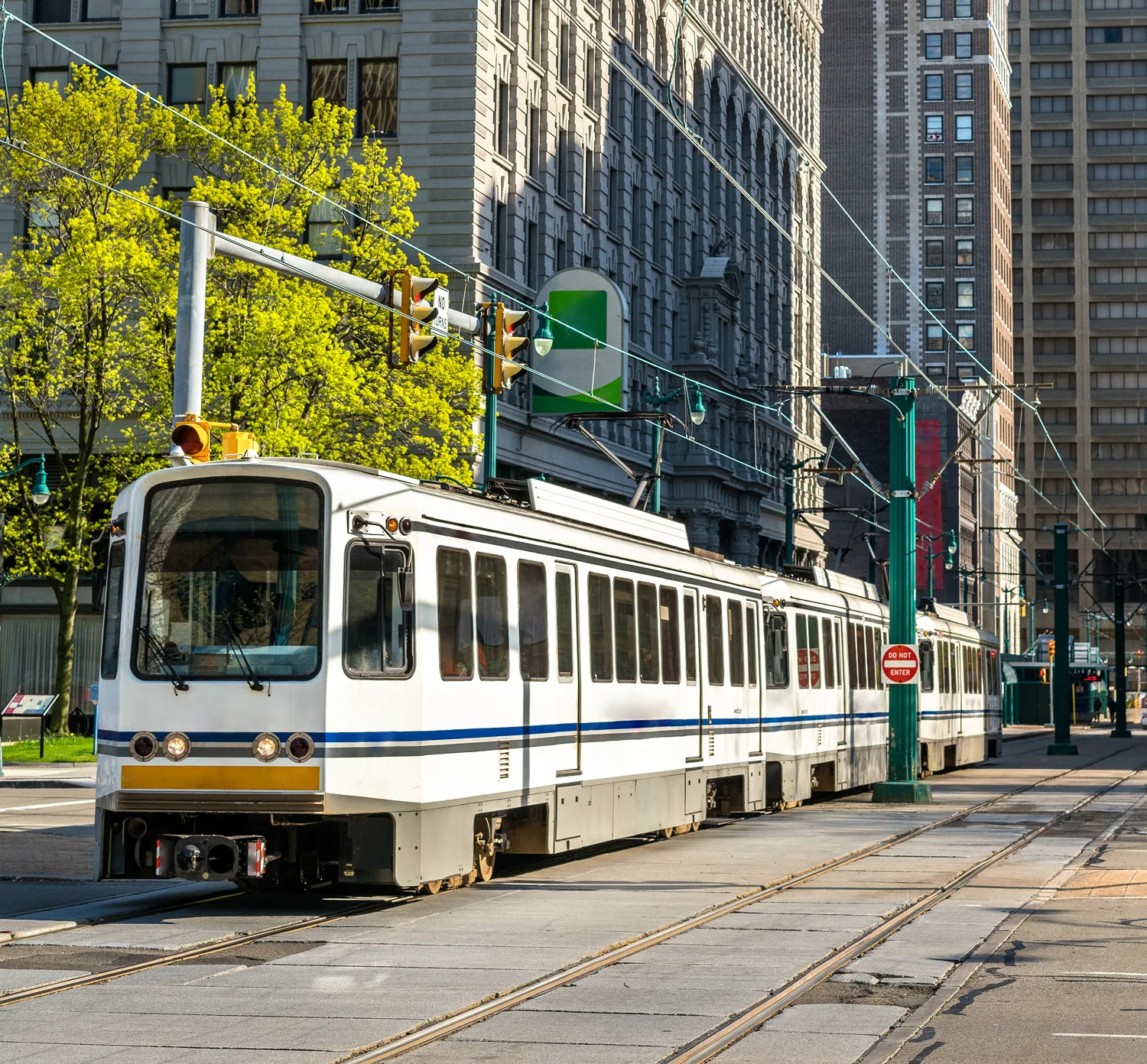 White and blue streetcar traveling on tracks along a city street with buildings and trees in the background.