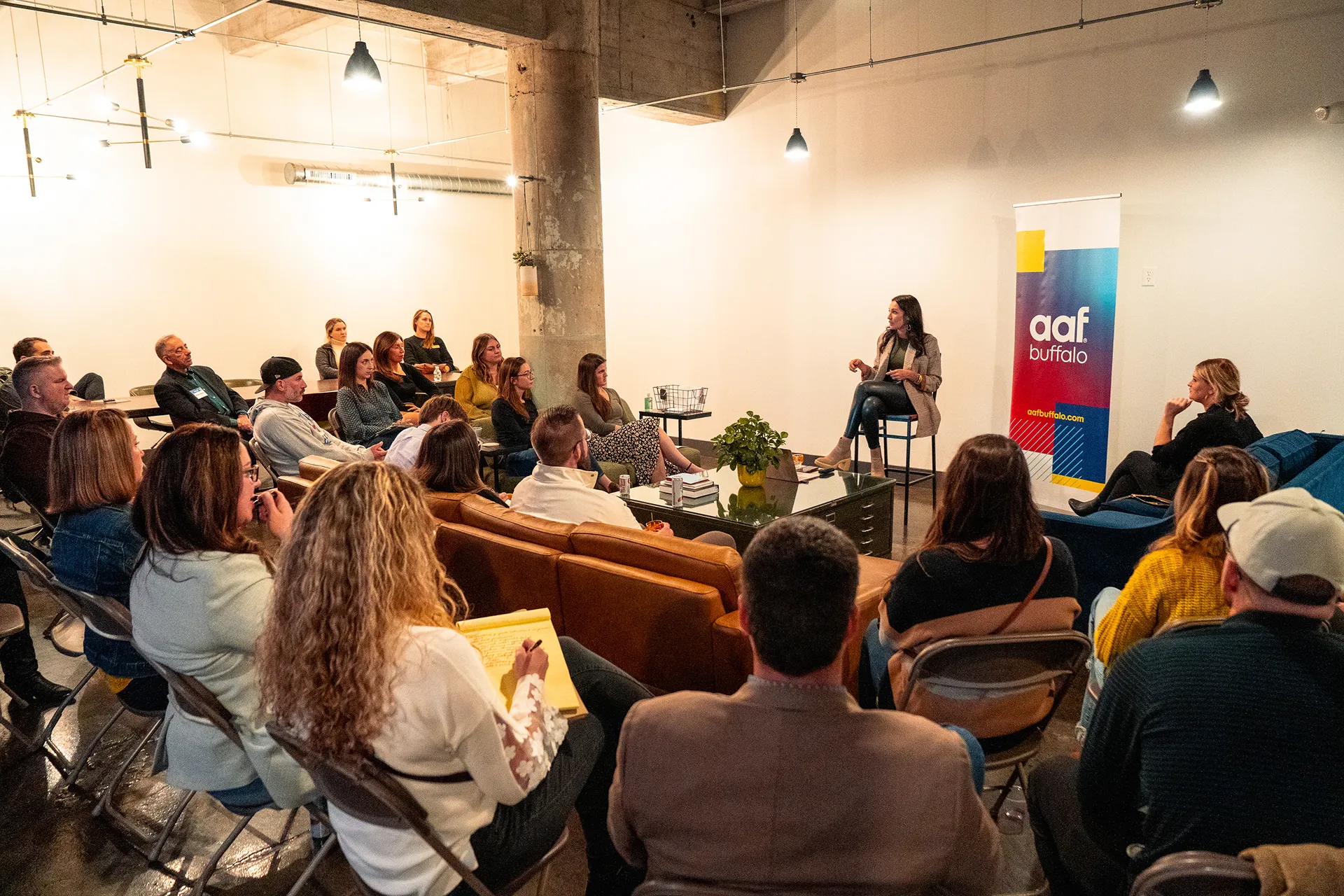 A group of people attentively listening to a woman speaking during an indoor seminar with an AAF Buffalo banner in the background.