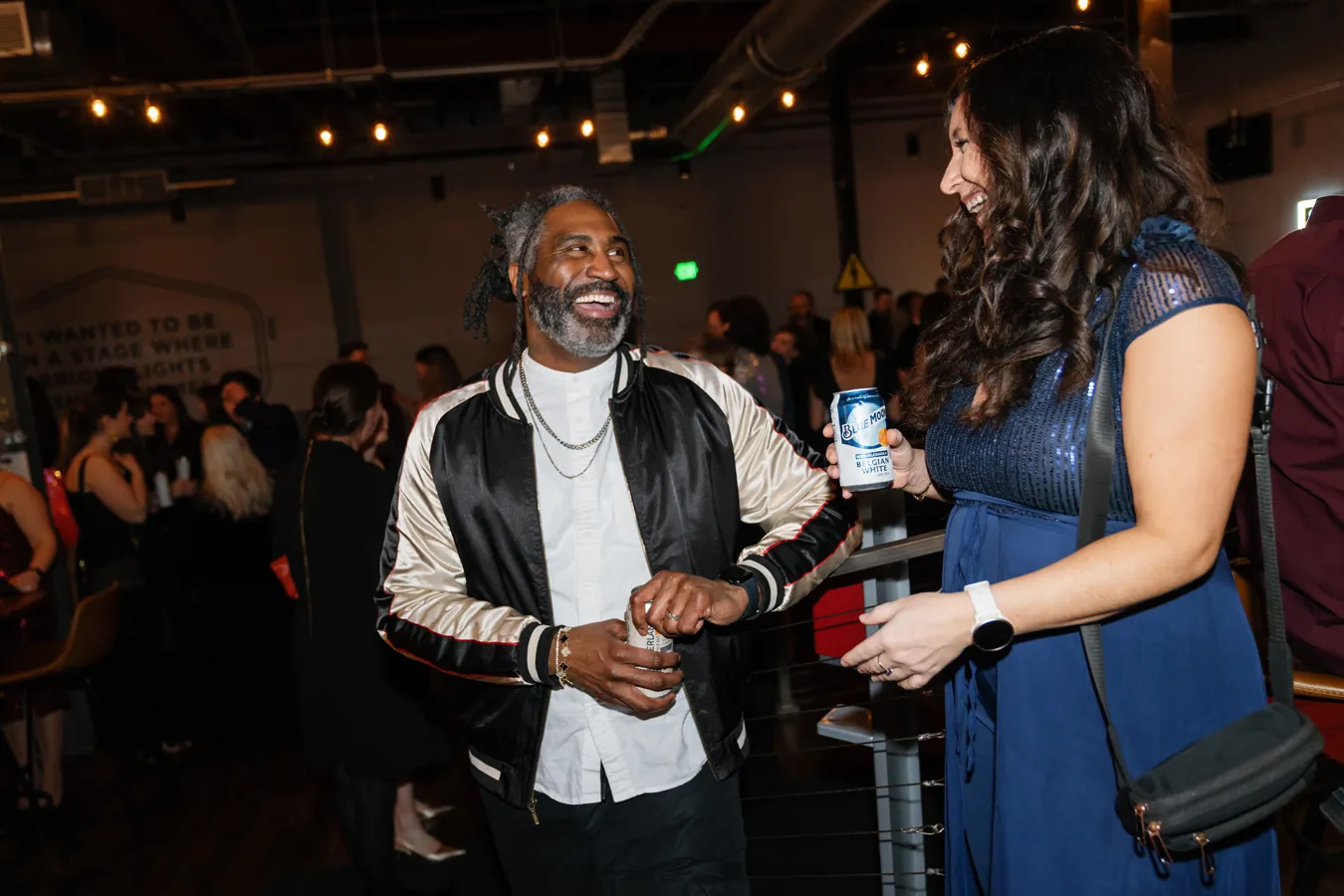 Man and woman smiling and holding canned drinks while socializing at a lively indoor event.