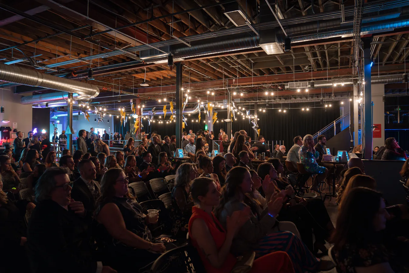Large audience seated and standing in an industrial-style event space with string lights and hanging decorations.