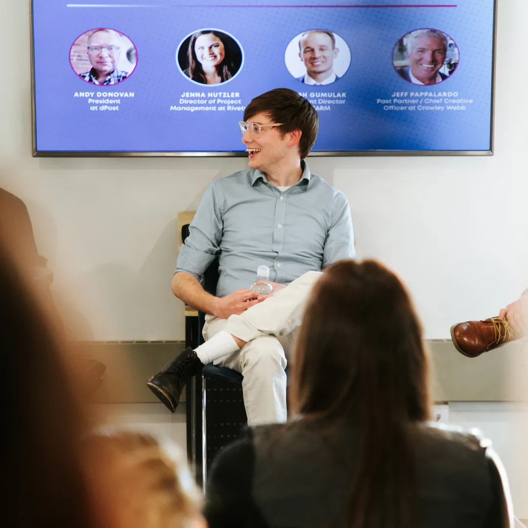 Young man wearing glasses and light shirt sits cross-legged on a chair, holding a water bottle and smiling in a meeting room with a screen displaying headshots and titles behind him.
