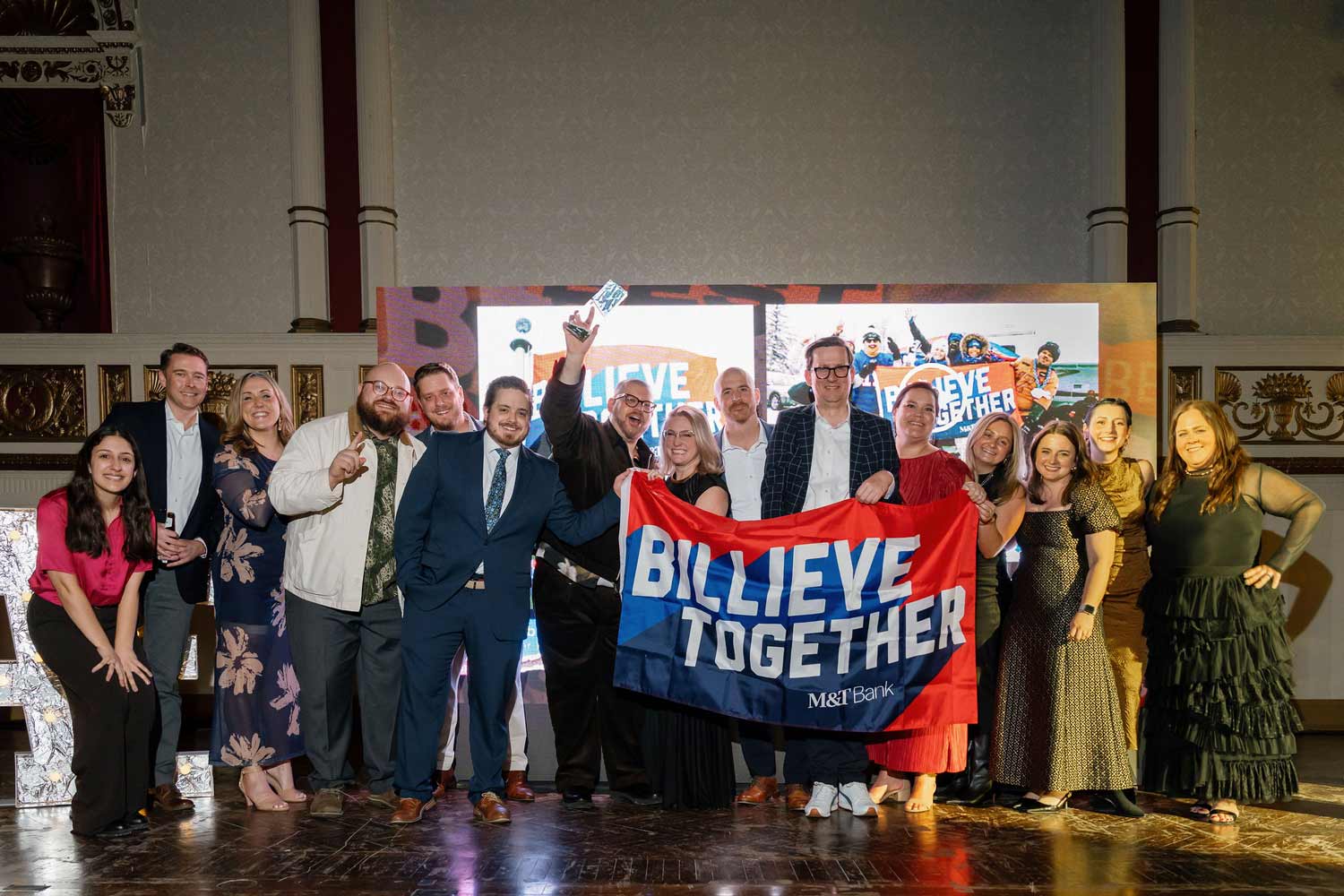 Group of diverse people, Crowley Webb, standing indoors holding a red and blue 'BILLIEVE TOGETHER M&T Bank' banner, smiling and celebrating.