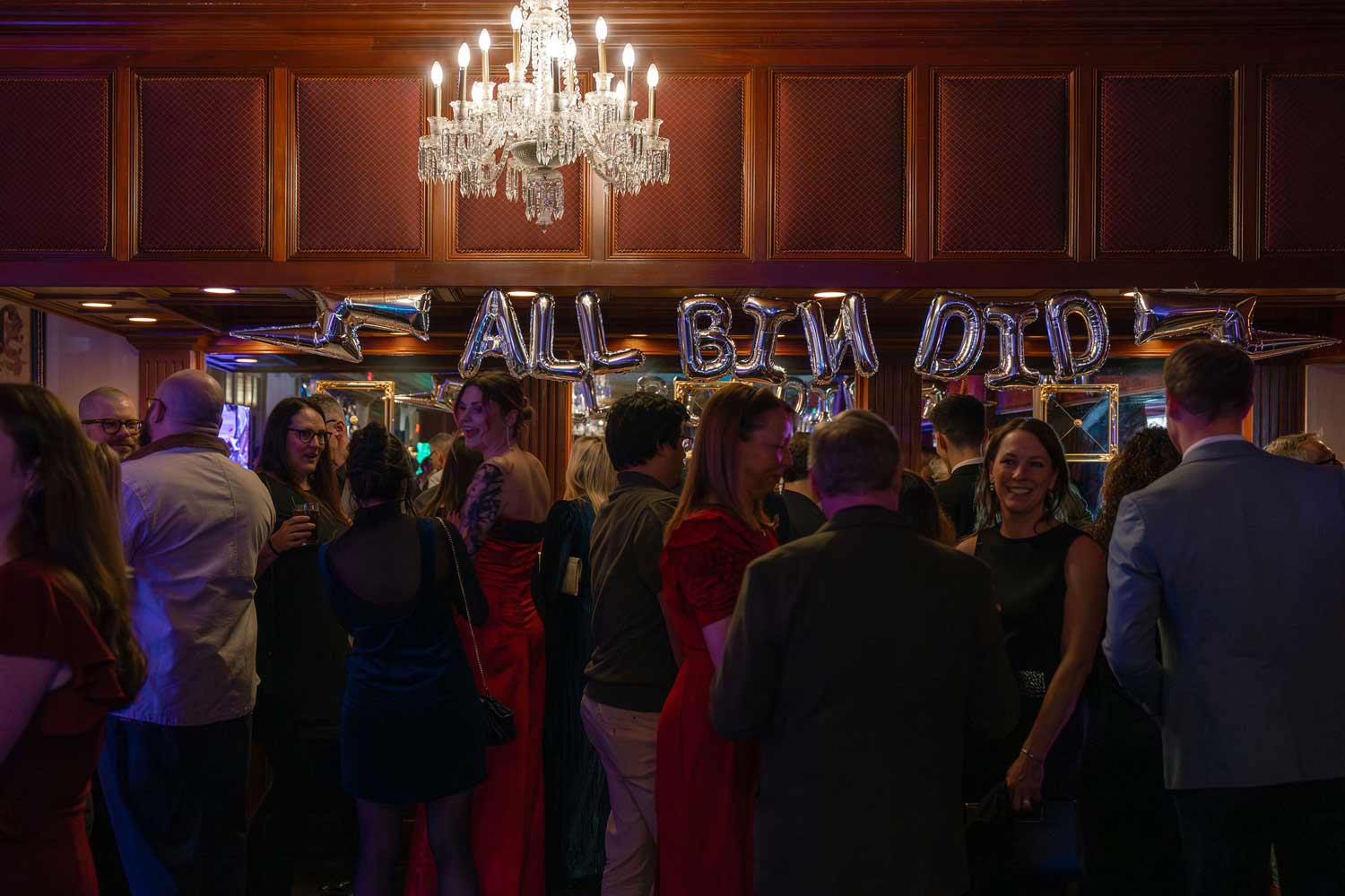 People dressed in formal attire socializing under balloon letters in a warmly lit room with a chandelier.
