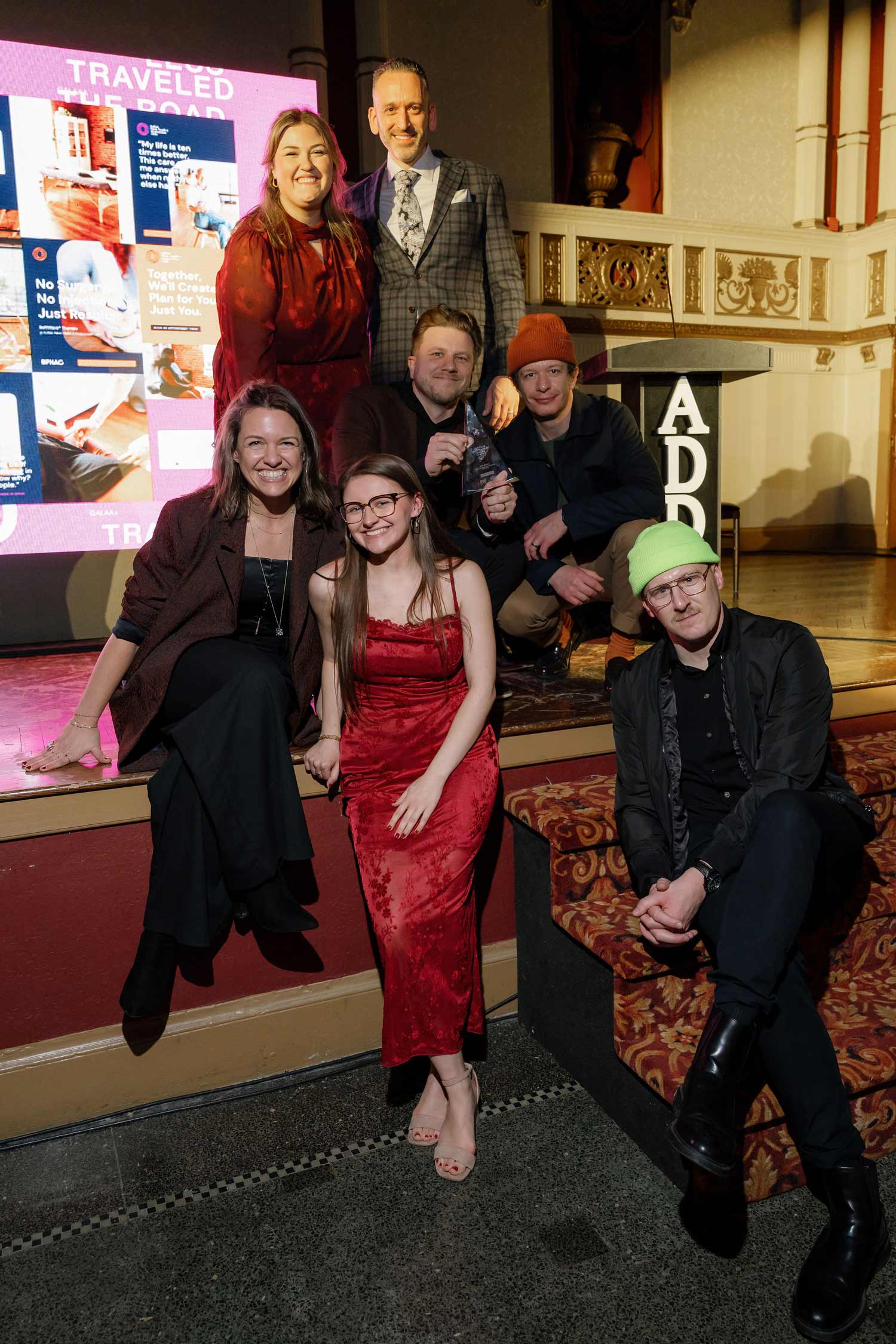 Group of seven people, Mr. Smith Agency, posing indoors at an event, with some seated on stage and carpeted steps, one holding a clear award trophy.