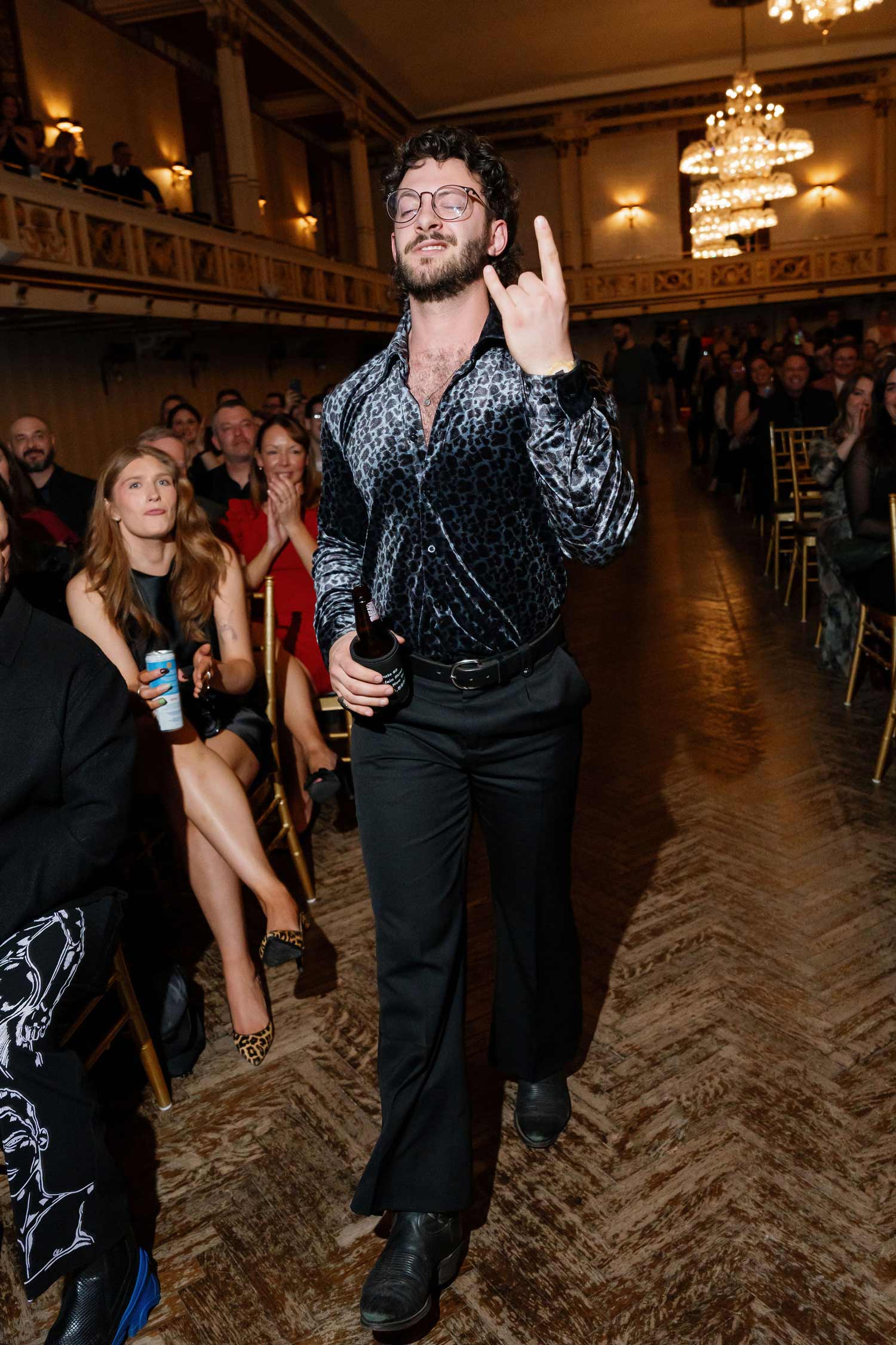 Man, Luke Hallick II, with curly hair and glasses holding a drink and gesturing rock sign while walking down an aisle in a fancy event hall with seated audience.