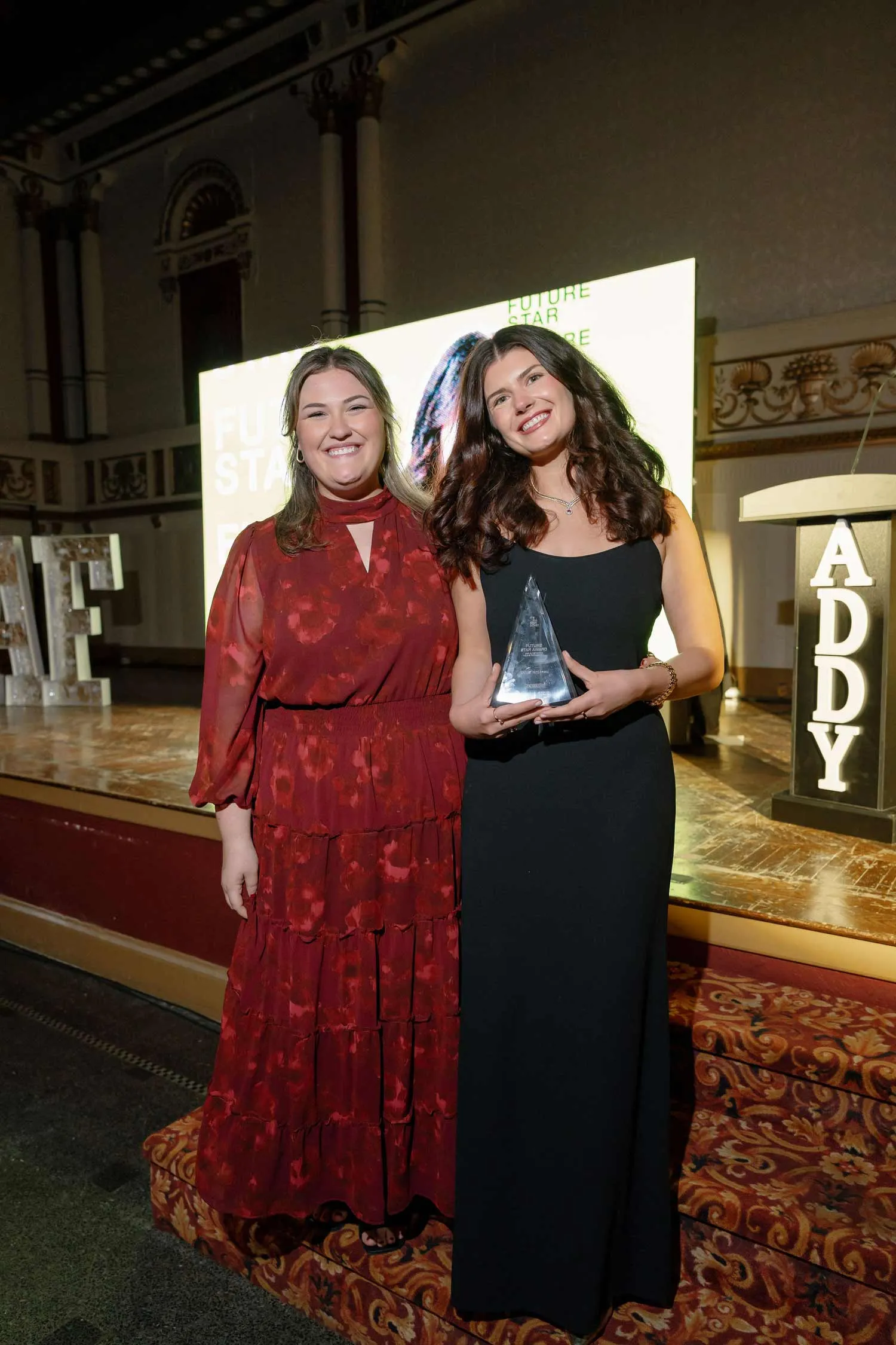 Two women, Lindsay Neilson and Hailey Rozanski, smiling on a stage, one holding an award, with illuminated ADDY letters in the background at an event.