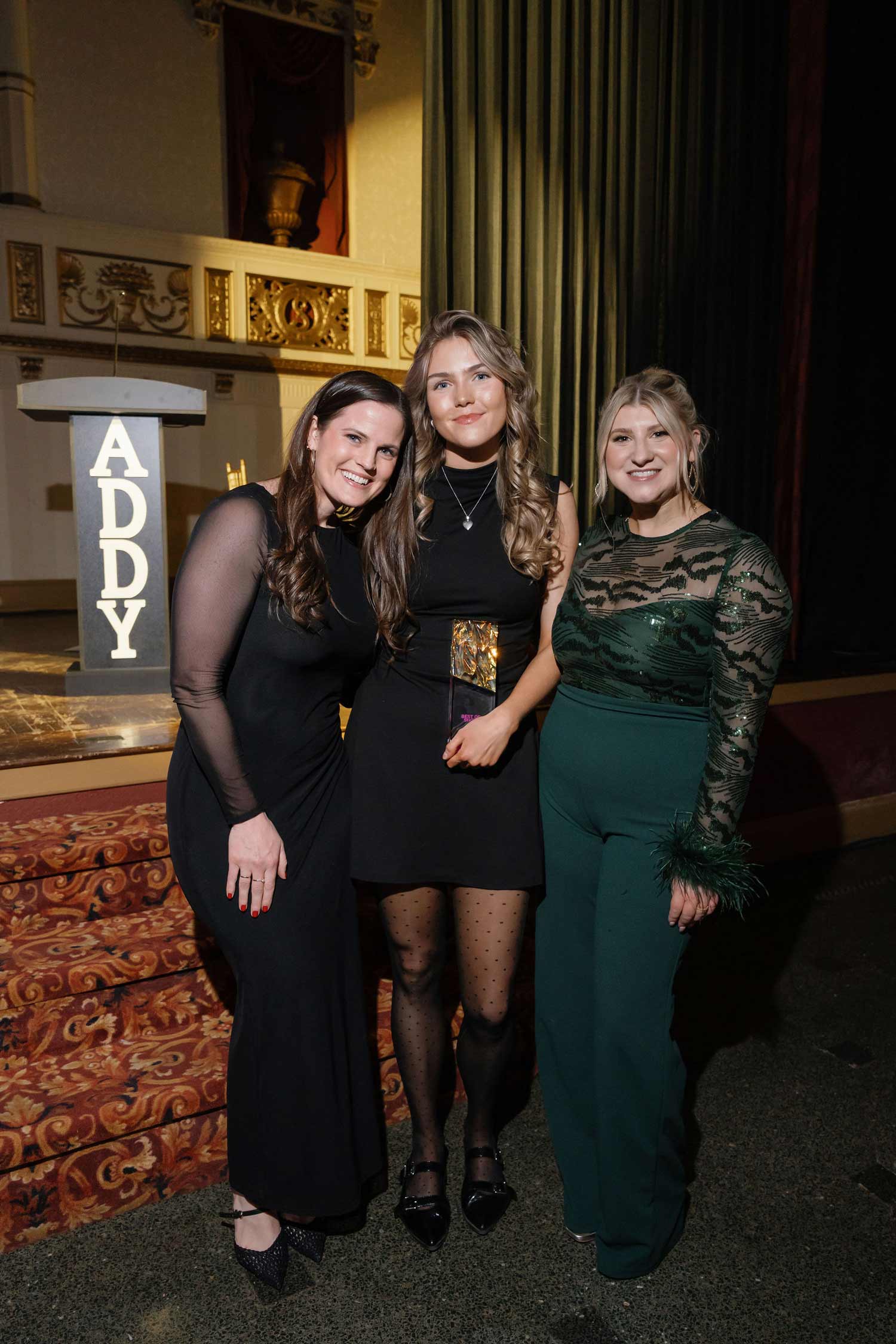Three women, Riveter, dressed in elegant evening attire smiling and posing together indoors near a podium with 'ADDY' letters.