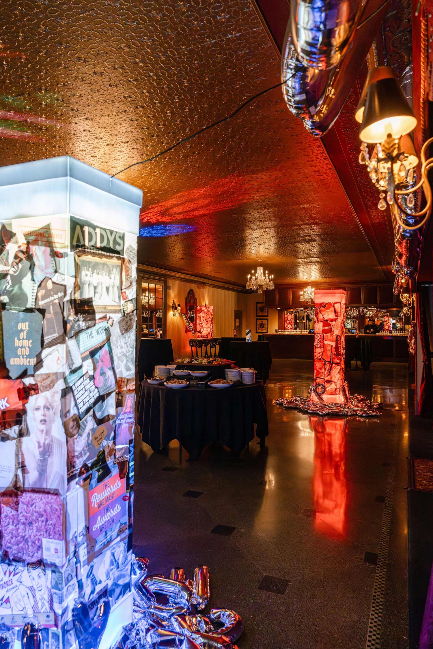 Interior of a dimly lit event space, The Statler Golden Ballroom, with two illuminated pillars decorated with collage images and silver balloons, round tables with black tablecloths, and chandeliers.