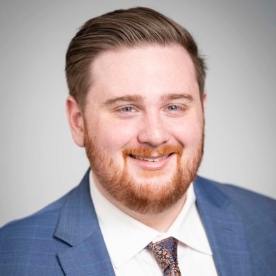 Smiling man with red hair and beard wearing a blue suit, white shirt, and patterned tie against a light gray background.