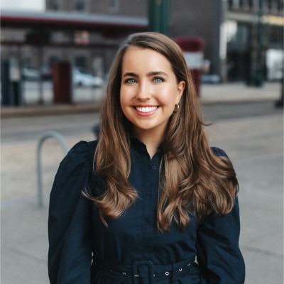 Smiling woman with long brown hair wearing a dark blue dress standing outdoors in an urban setting.