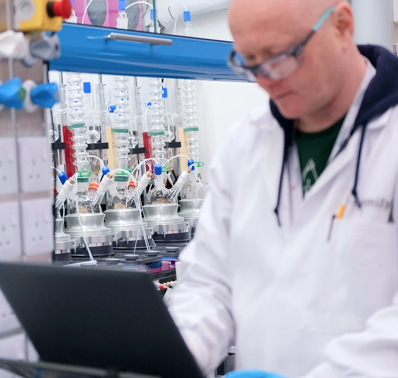 A scientist works at a laptop with a robotic chemistry platform in the background