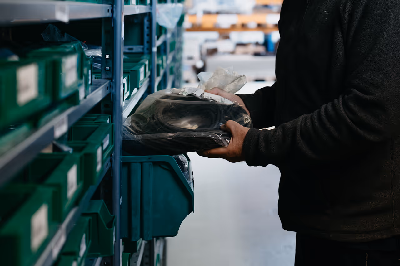 Eine Person hält verpackte Teile in einem Lager mit grünen Kunststoffbehältern an Metallregalen.