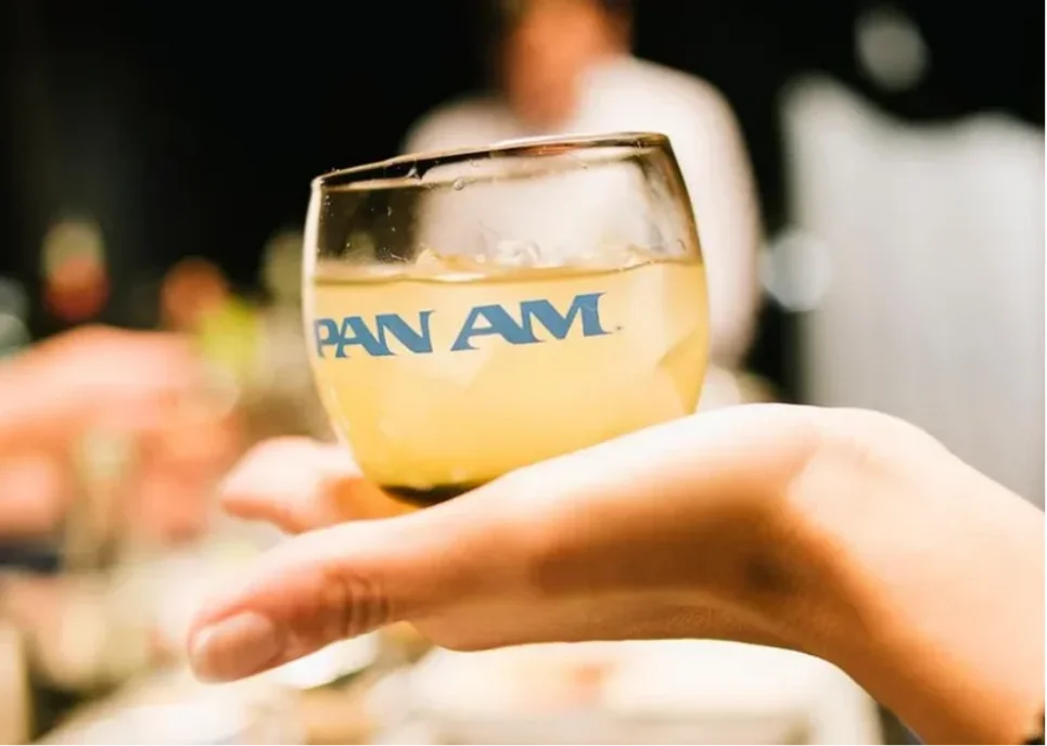 A hand holds a glass bearing the Pan Am logo, capturing the signature elegance of in-flight service.