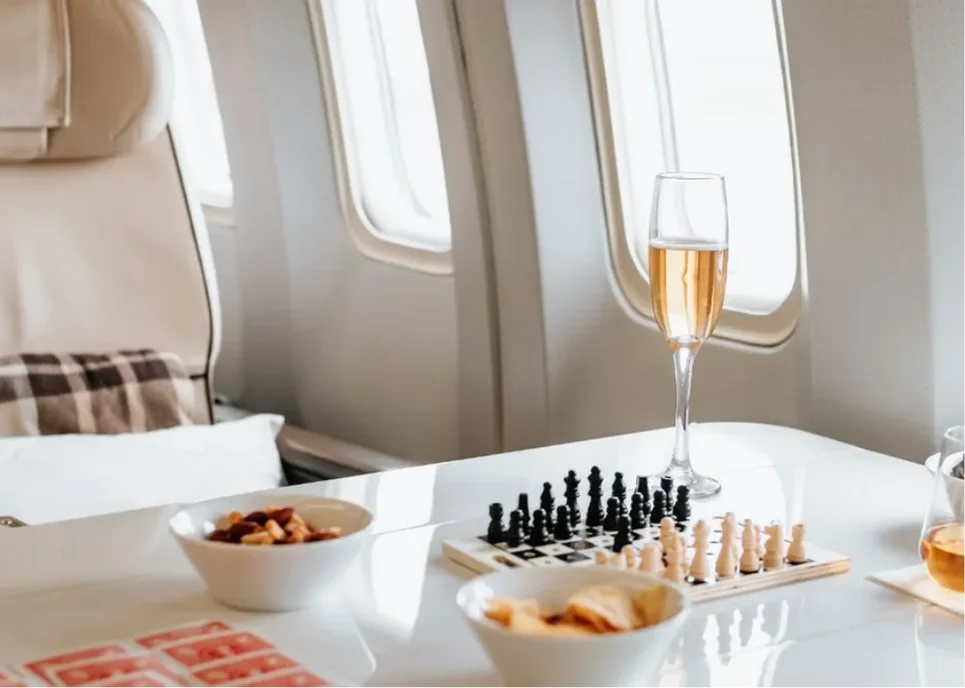 A glass of champagne, a travel chess set, and small dishes rest on a table beside a plush Pan Am seat mid-flight.