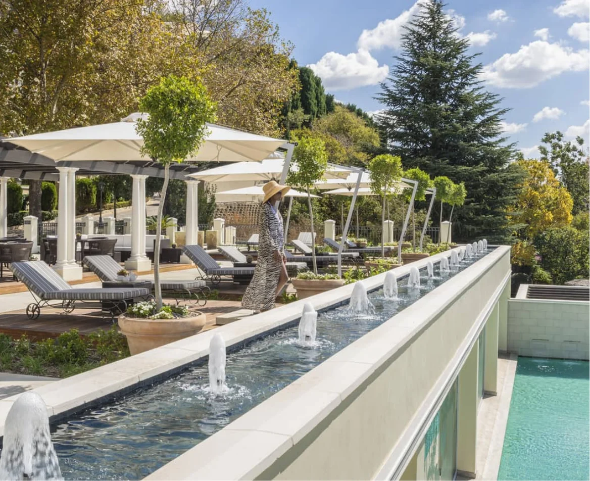 Guest walks beside a sunlit fountain and shaded lounge chairs at a luxury hotel terrace surrounded by trees.