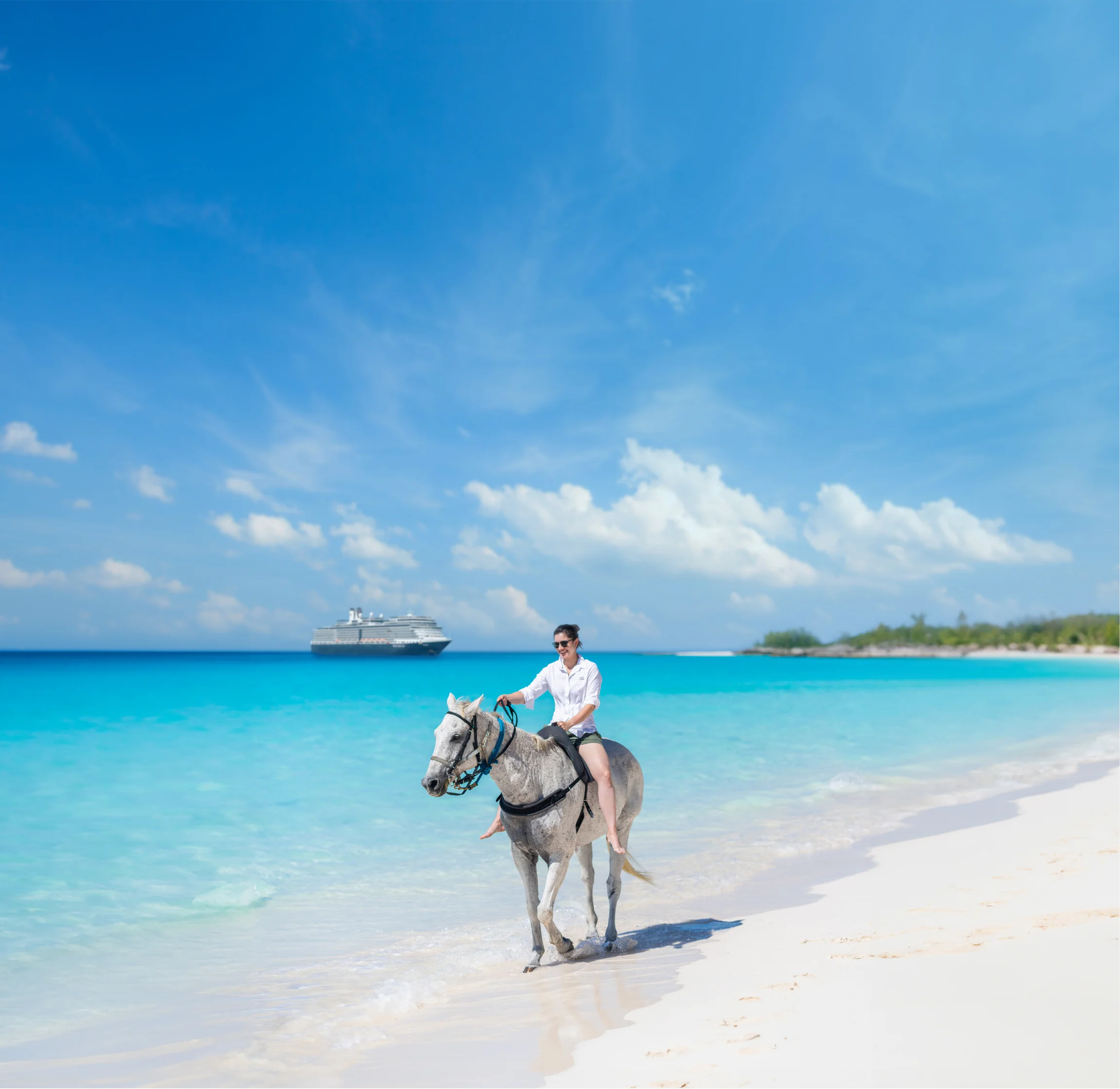 Woman riding a white horse along the shoreline of a tropical beach with turquoise water and a cruise ship in the distance.