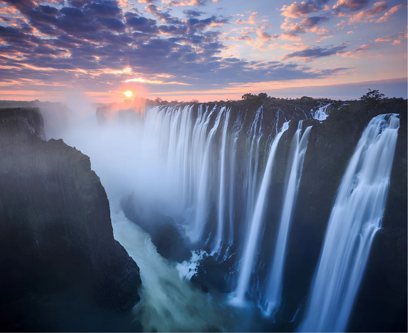 Sunset over a wide, misty waterfall cascading down rocky cliffs with scattered clouds in the sky.