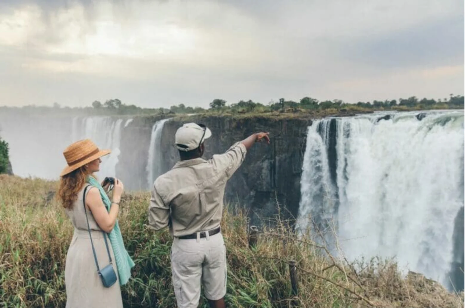Two tourists near tall, wide waterfall with mist, one pointing and the other holding binoculars.