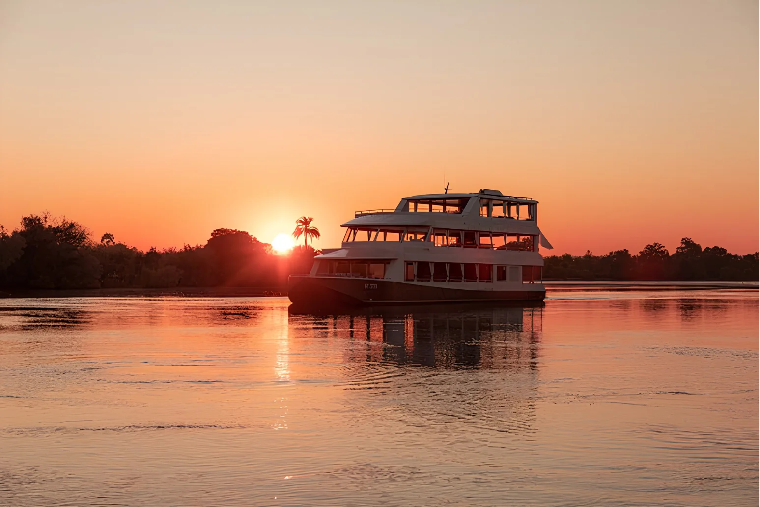 Boat floating on calm water at sunset with trees and palm silhouette on the horizon.