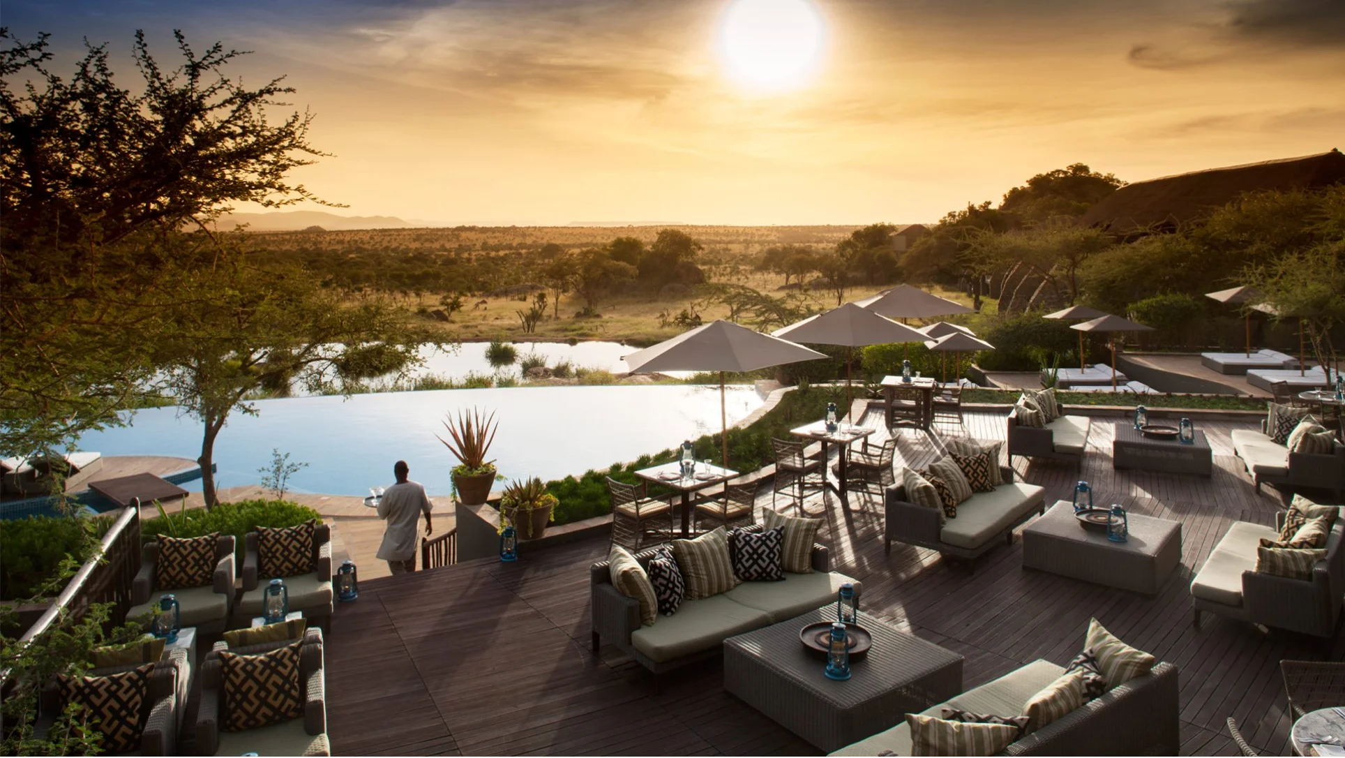 Outdoor lounge and dining area with cushioned seating and umbrellas near an infinity pool overlooking a savannah at sunset.