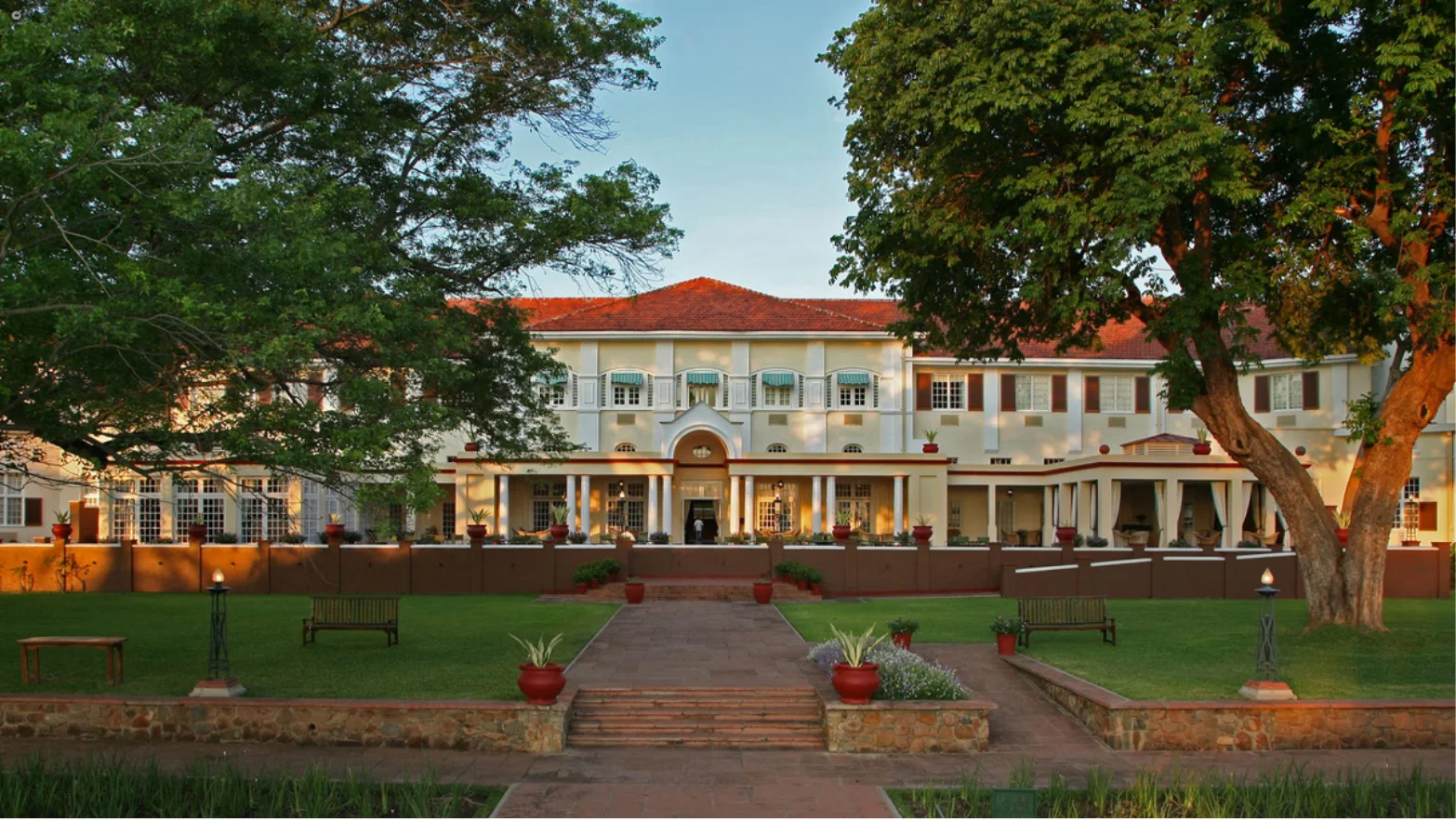 Large colonial-style building with red roof and white exterior, surrounded by green lawn, trees, and benches.