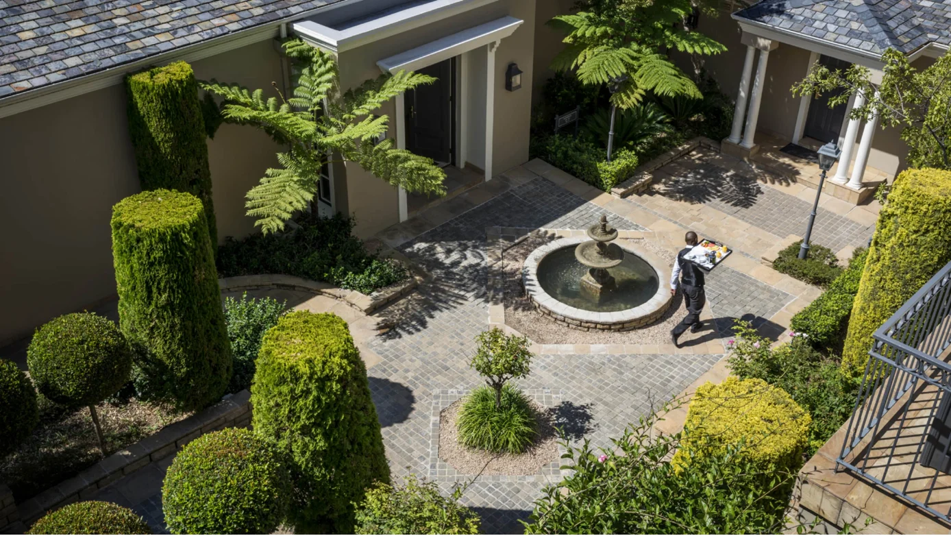 A courtyard with trimmed green bushes, a round stone fountain in the center, and a person carrying a tray walking on a cobblestone path.