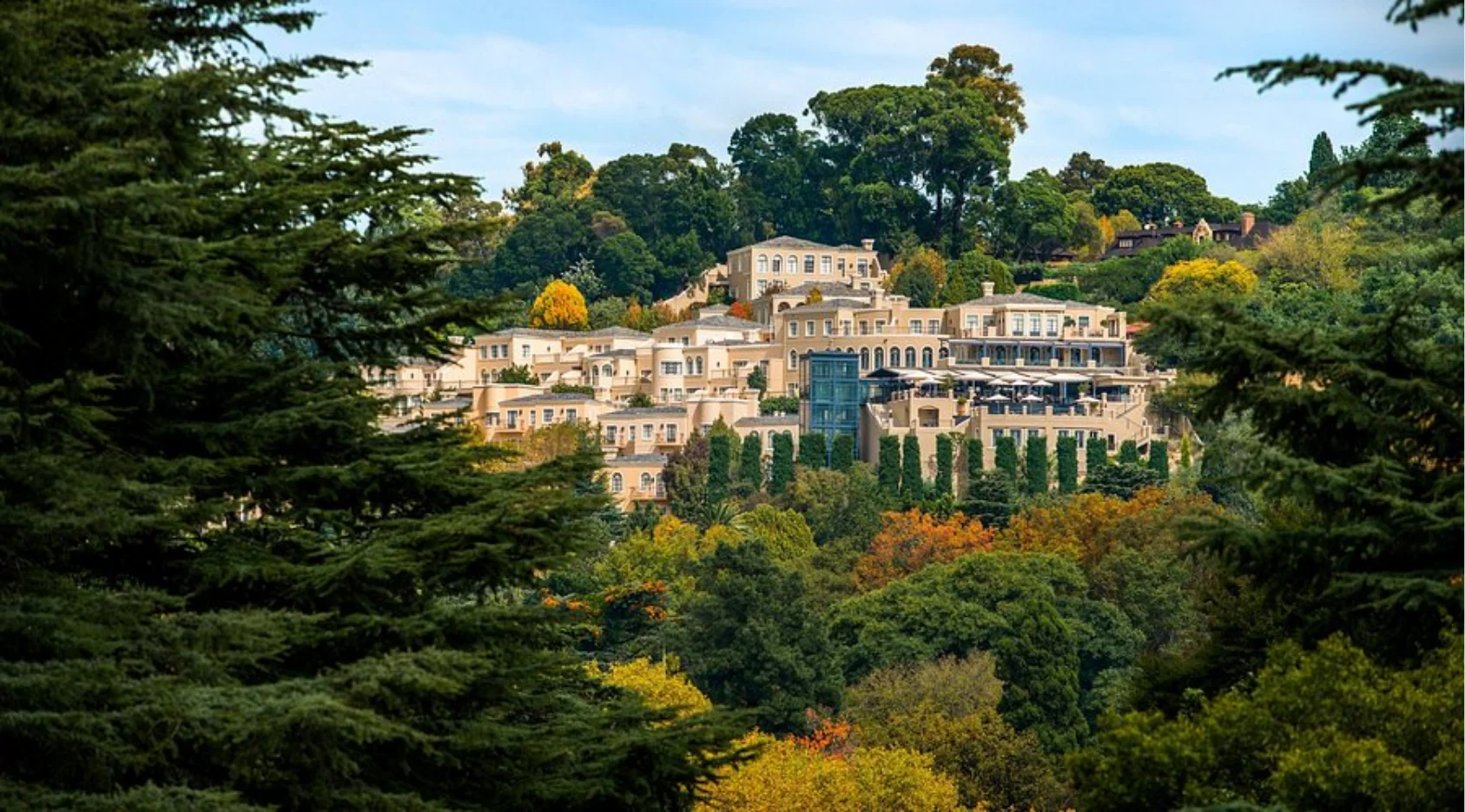 Large beige multi-story building complex surrounded by dense green and autumn-colored trees on a hillside.