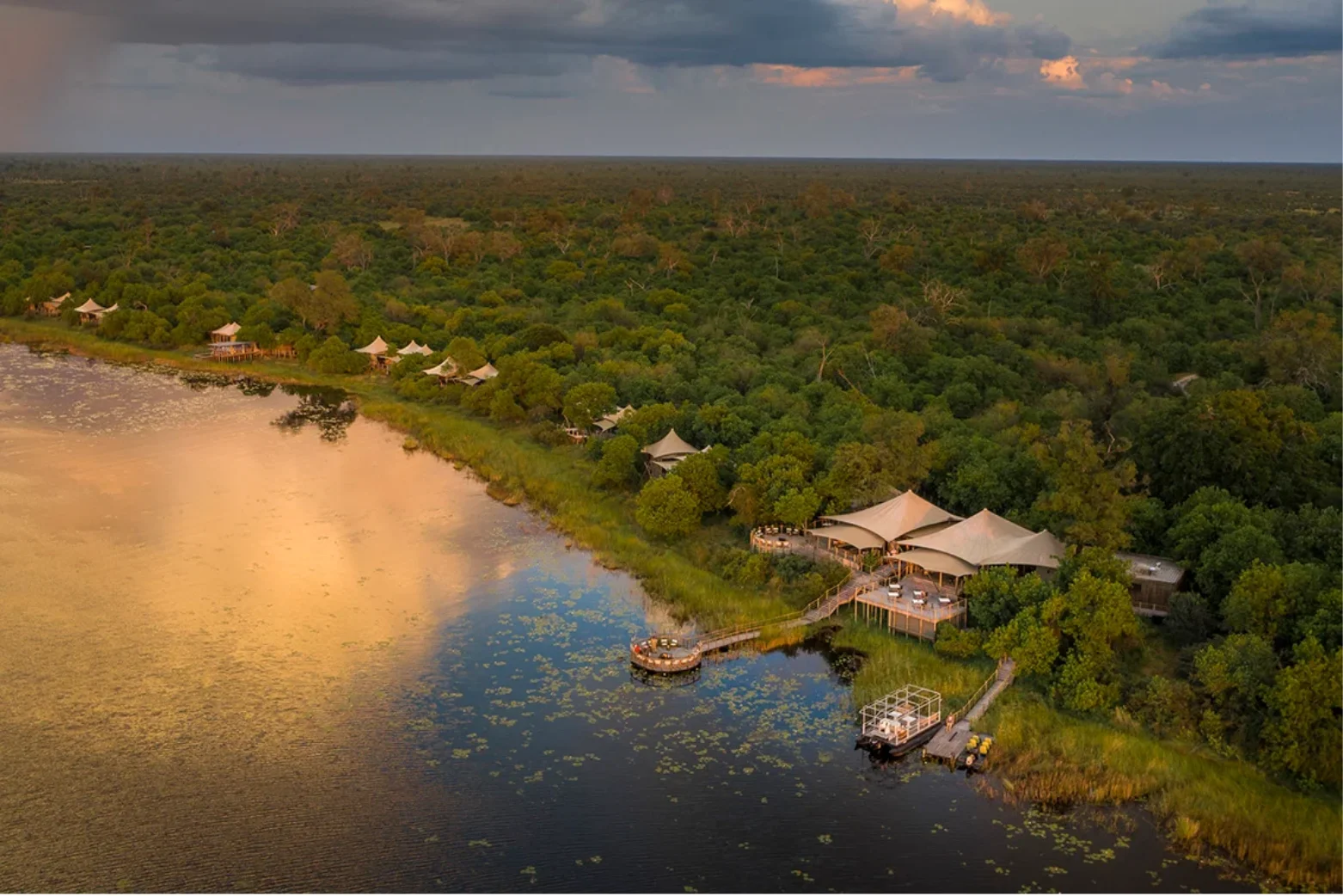 Aerial view of a lakeside safari lodge with tents surrounded by dense forest under a cloudy sky at sunset.