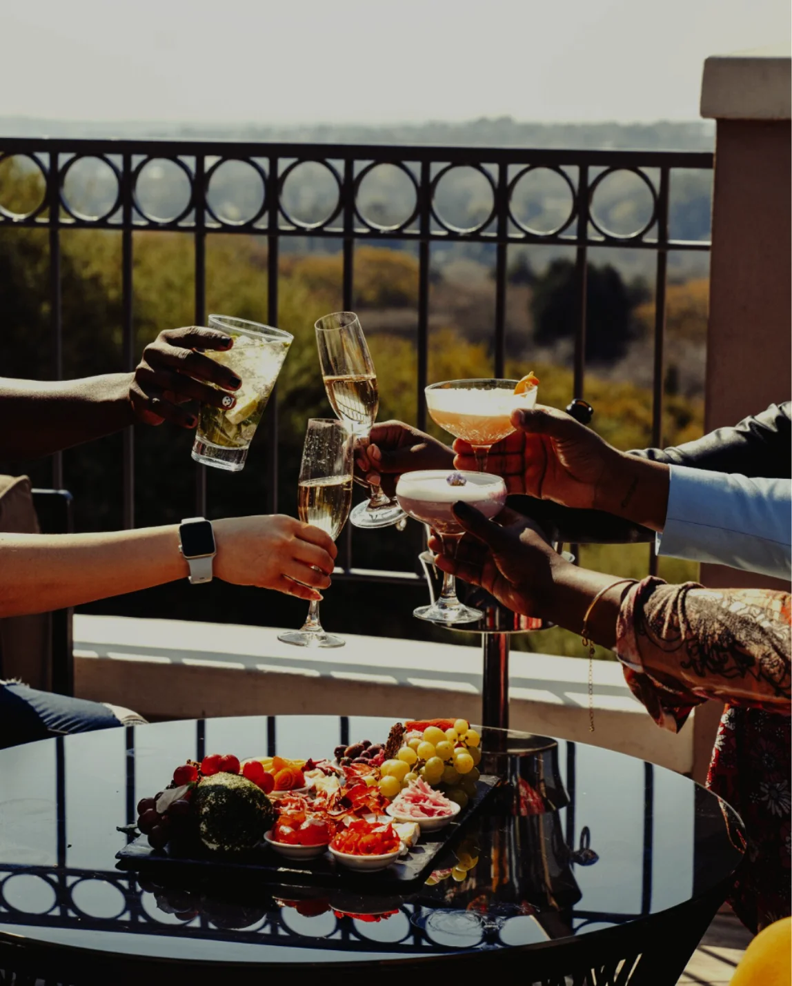 Group of people clinking glasses of cocktails and champagne above a table with assorted appetizers on a terrace with a scenic view.