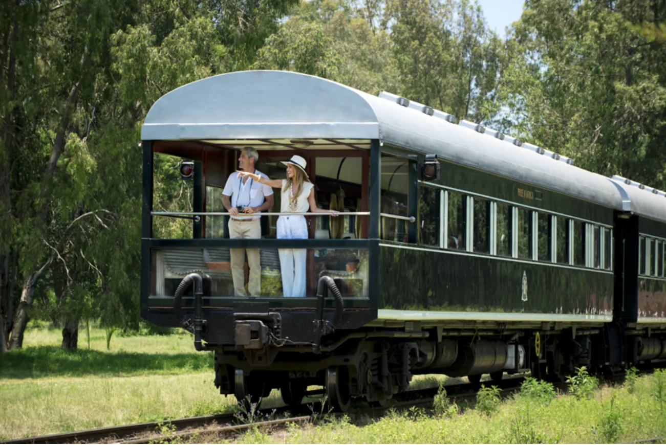 Two people standing and enjoying the view on the rear outdoor platform of a vintage dark green passenger train carriage in a green natural setting.