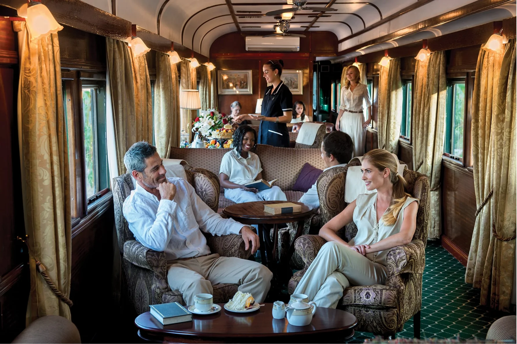 Elegant vintage train car lounge with passengers seated in patterned armchairs, a server offering refreshments, and warm interior lighting.