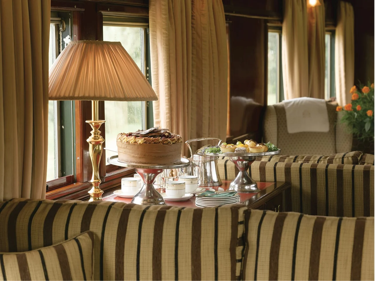 Cozy train lounge with striped upholstered seats, a table with cake, pastries, tea set, and a brass lamp by the window.