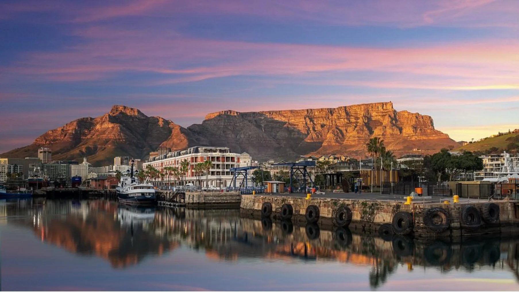 Sunlit Table Mountain and city waterfront with boats reflected in calm water during sunset.