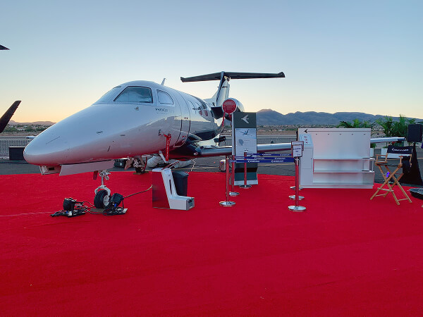 Modern small private jet displayed on red carpet with mountains in the background at sunset.
