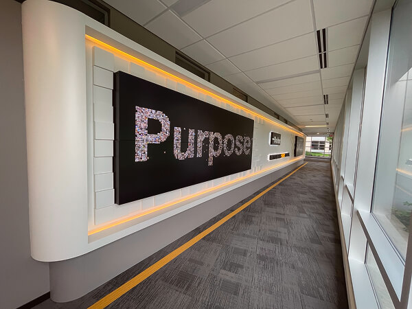 Modern hallway with a large black wall panel displaying the word 'Purpose' made up of small photos, illuminated by soft yellow backlighting.
