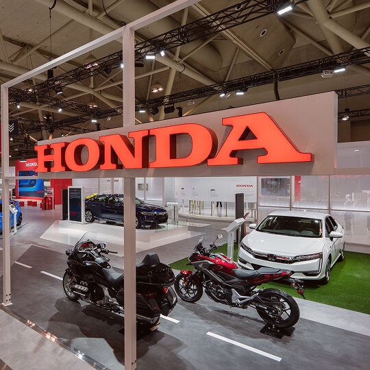 Honda showroom display featuring two motorcycles in the foreground and a white sedan in the background under a large red Honda sign.