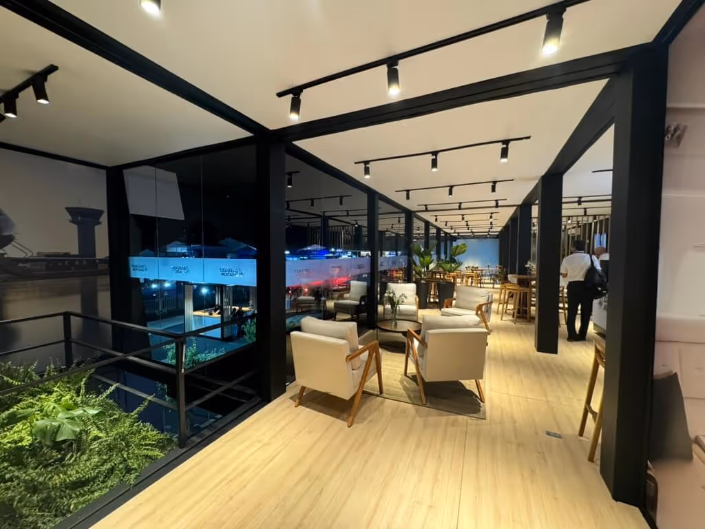 Modern indoor lounge area with beige chairs, wooden floor, black beams, large glass windows, and two people near a counter.