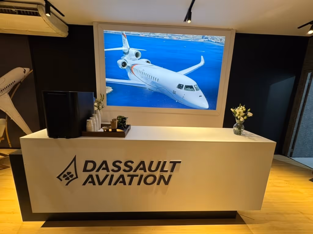 Dassault Aviation reception desk with logo, a model airplane on the left wall, and a large screen displaying a white private jet flying over blue water.