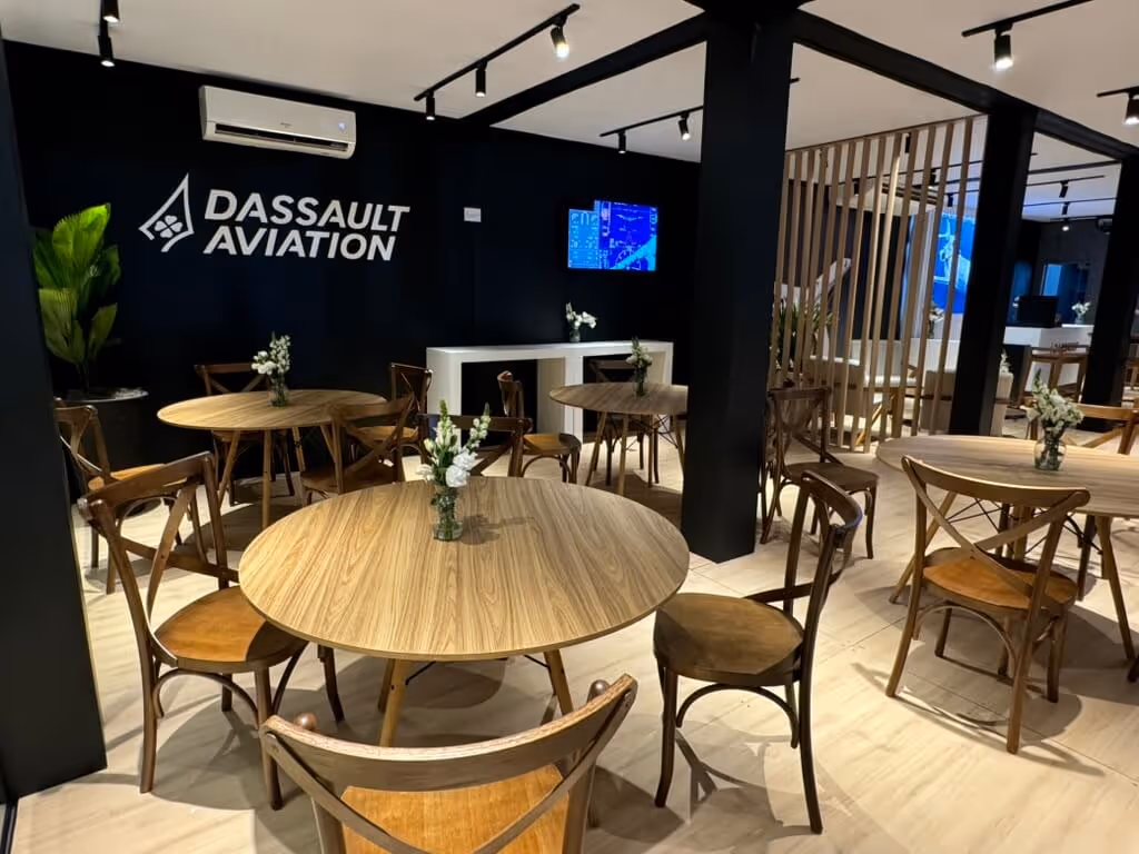 Modern office lounge with round wooden tables, wooden chairs, small flower vases, and a black wall with Dassault Aviation logo.