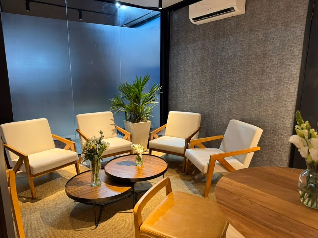 Modern seating area with four beige armchairs, round wooden coffee tables with flower vases, a potted plant, and an air conditioner on the wall.