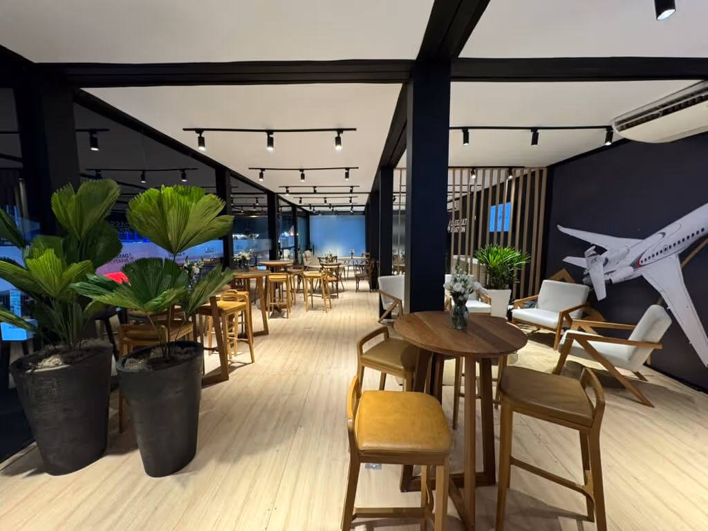 Modern indoor lounge area with wooden chairs and tables, potted plants, and a large airplane model mounted on a dark wall.