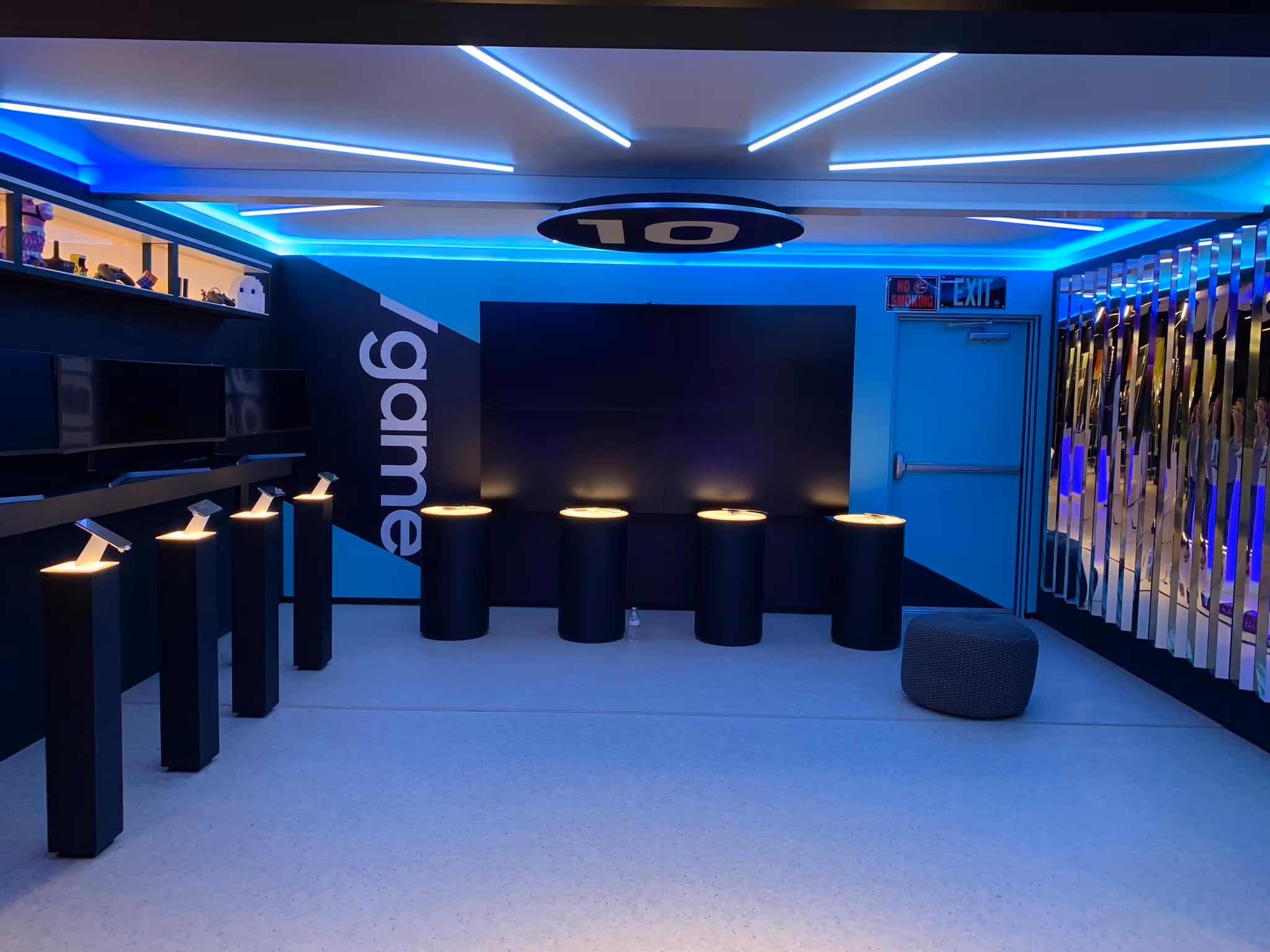 Futuristic gaming room with blue LED lights, five illuminated cylindrical stools, multiple screens, and a padded gray ottoman.
